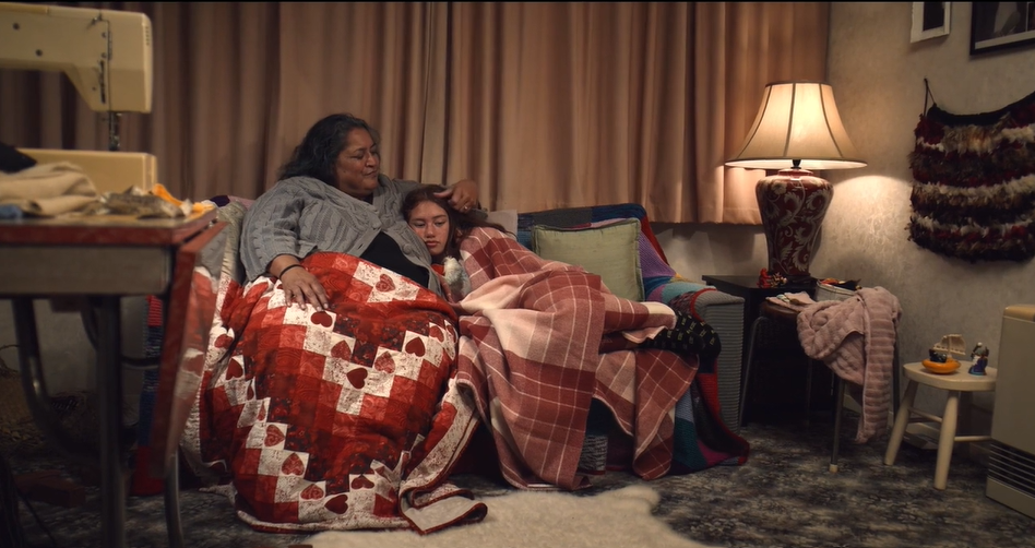 An older woman and a young girl lie cuddled up on a sofa, both covered in bright blankets.  