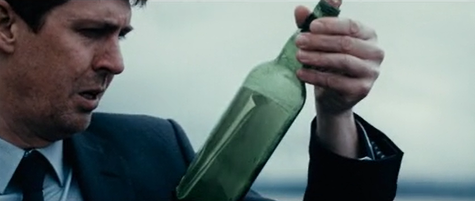 A man in a suit holds a green bottle up closely to his eyes