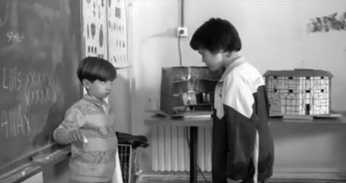 Black and white image. A small boy stands with his back to a blackboard, looking up at a larger boy who is looming over him. 