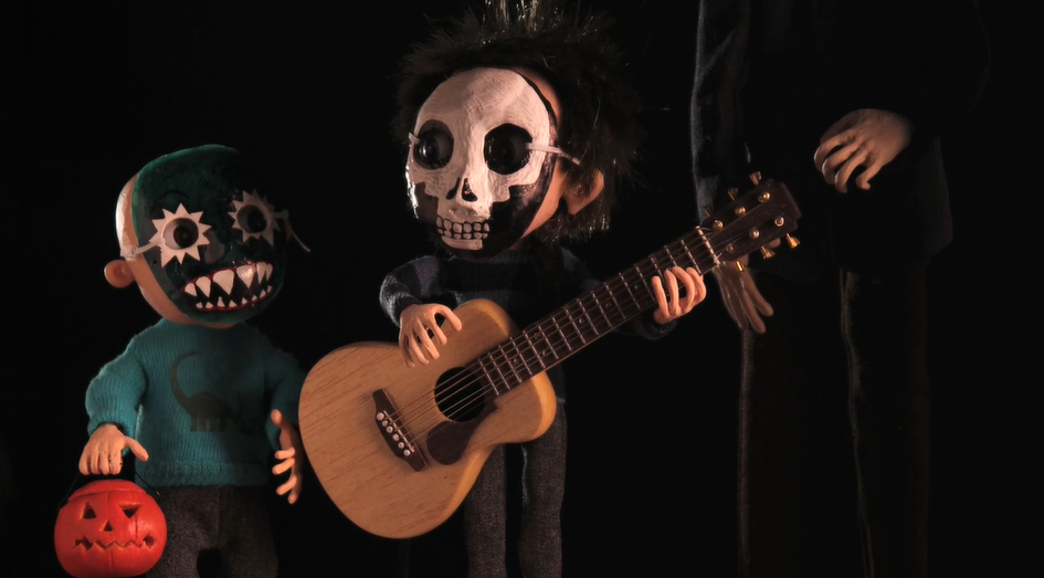 Two small boys in Hallowe&#039;en masks stand next to an adult, whose legs we can only see.  One boys holds a small pumpkin with a face carved in it, one holds a guitar.