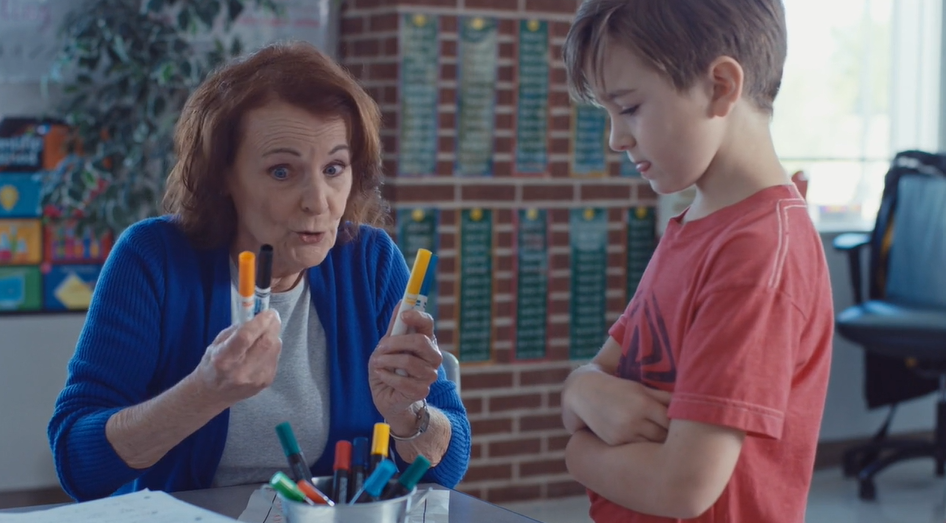 A middle-aged female teacher holds a few colouring pens up to a young boy who is standing with his arms crossed, looking disinterested.