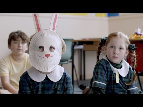 Three children sitting in a classroom.  One is wearing a bunny mask made out of a paper plate.